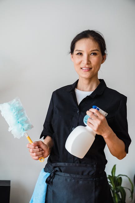A female cleaner holding a dusting brush in her right hand and a spray bottle in her left hand, standing indoors against a plain light gray wall. She is dressed in a black uniform with a white inner s