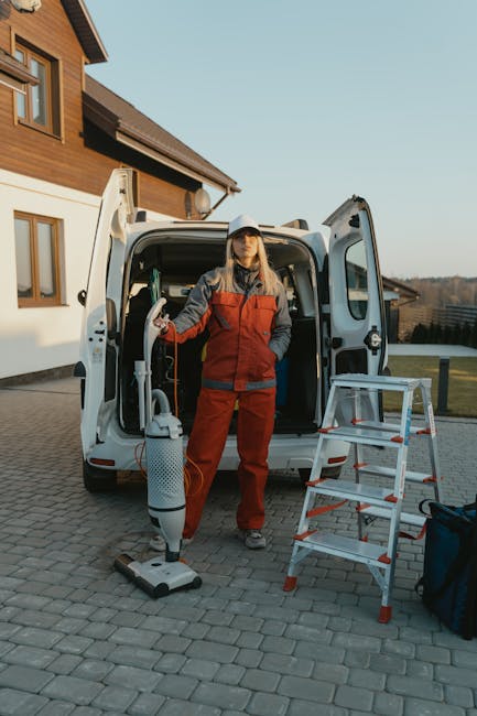 Two professional cleaners wearing red uniforms are performing surface cleaning in a modern living room, using vacuum cleaners on the tiled floor. The room features a contemporary grey sofa with decora