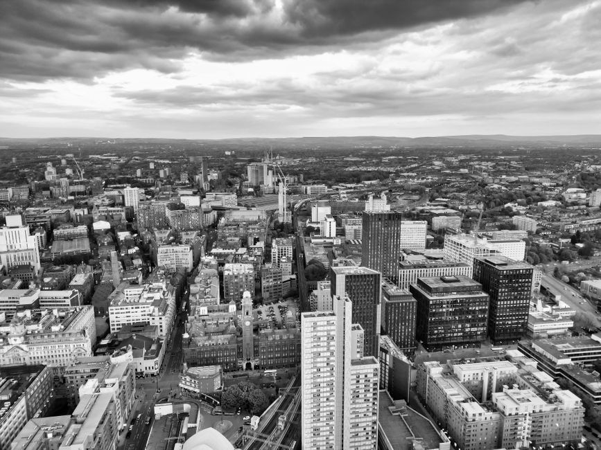 A black and white aerial view of a densely built cityscape featuring high-rise office buildings, residential apartments, and commercial structures. The scene shows a mix of modern glass and traditiona