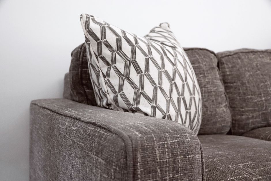 A person wearing a black leather jacket is vacuuming a yellow and white patterned cushion on a dark fabric sofa in a living room. The scene highlights surface cleaning and deep cleansing of upholstery